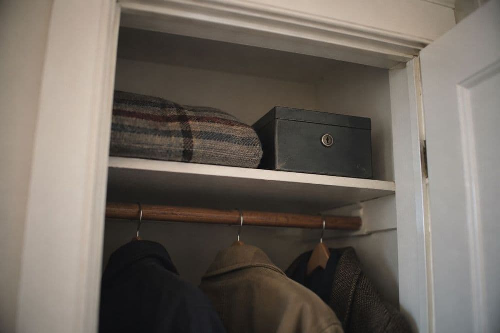 A small closed metal lockbox on the top shelf of a hall closet, well above a row of hanging coats, in soft daylight.
