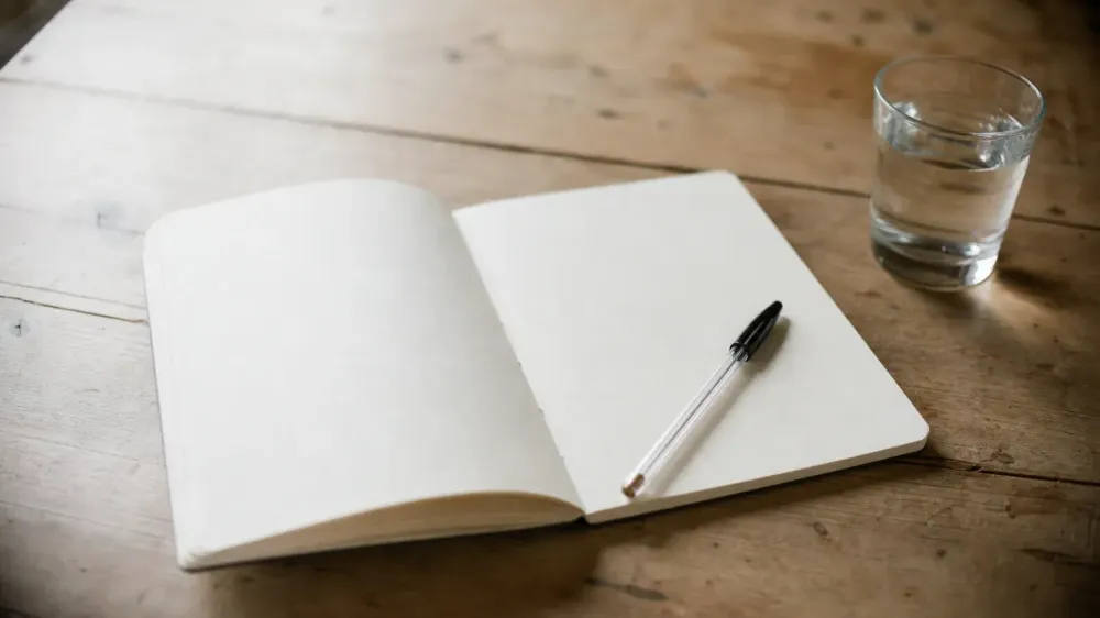 An open blank paper notebook and a pen beside a glass of water on a worn wooden kitchen table in morning light.