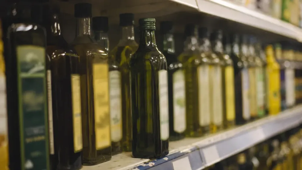 A grocery shelf of cooking oil bottles seen slightly from the side in ordinary store lighting, one dark green bottle pulled forward from the row, labels turned away and unreadable.