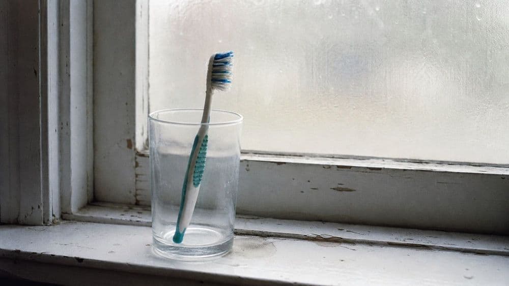 A worn toothbrush standing in a plain glass on a bathroom windowsill in flat daylight, with a chipped painted sill and a slightly steamed window behind it.