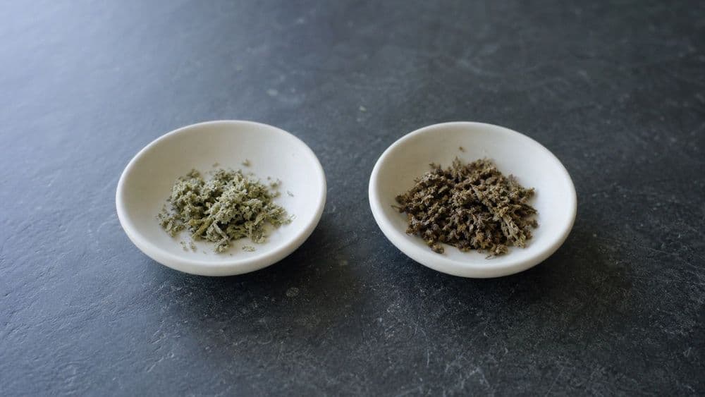 Two small plain dishes side by side on a slate counter, one holding pale raw plant material and the other holding the same material darkened after processing.