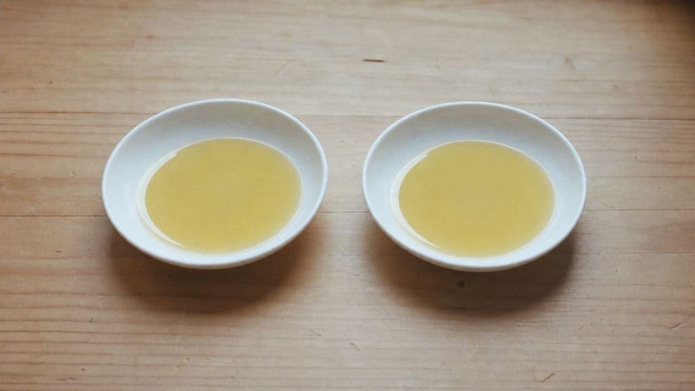 Two identical small white ceramic dishes side by side on a pale wooden table, each holding the same thin pool of pale golden oil, lit by soft daylight from one side.